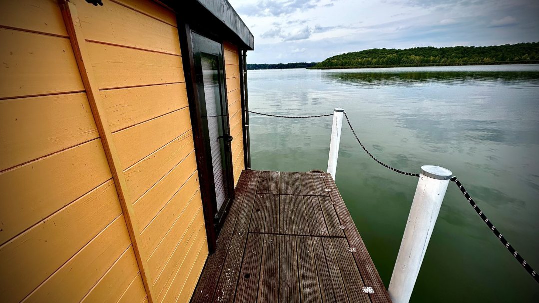 Holzsteg an einem Gewässer mit einer Gelben Seitenwand im Hintergrund.