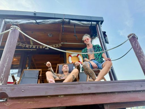 Zwei Personen entspannen auf einer Plattform, umgeben von Ketten und Blick aufs Wasser.