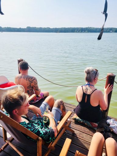 Menschen sitzen auf einer Holzterrasse am Wasser und genießen den Ausblick.