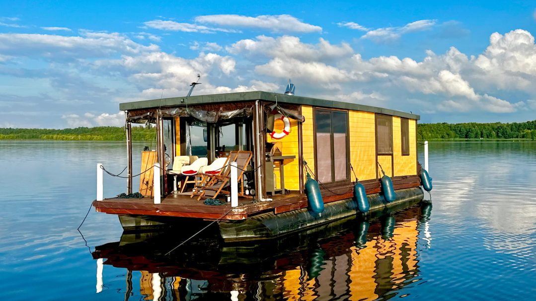 Bunte Hausboot auf ruhigem Wasser, umgeben von grünem Ufer und blauem Himmel mit Wolken.