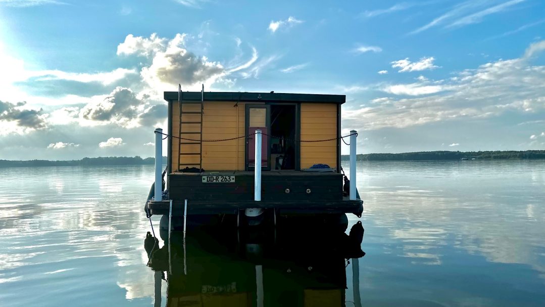 Ein Hausboot auf ruhigem Wasser, umgeben von blauem Himmel und Wolken.